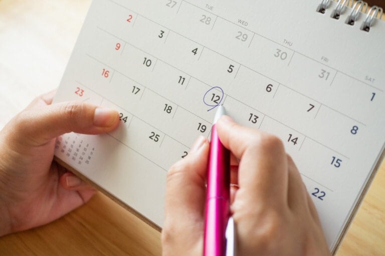 A person is holding a calendar while marking the date, the 12th, with a pink pen. The calendar shows the days of the month with numbers written in different colors—black, red, and blue—on a light background, ideal for tracking natural family planning.
