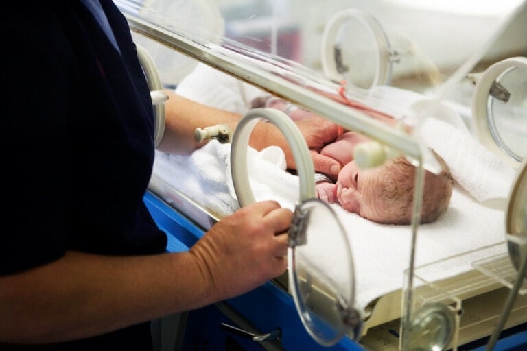 A newborn baby lies in an incubator with its eyes closed. An adult, likely one of the dedicated NICU nurses, stands beside the incubator with one hand on the transparent cover and the other inside, gently touching the baby. The scene appears to be in a medical facility.