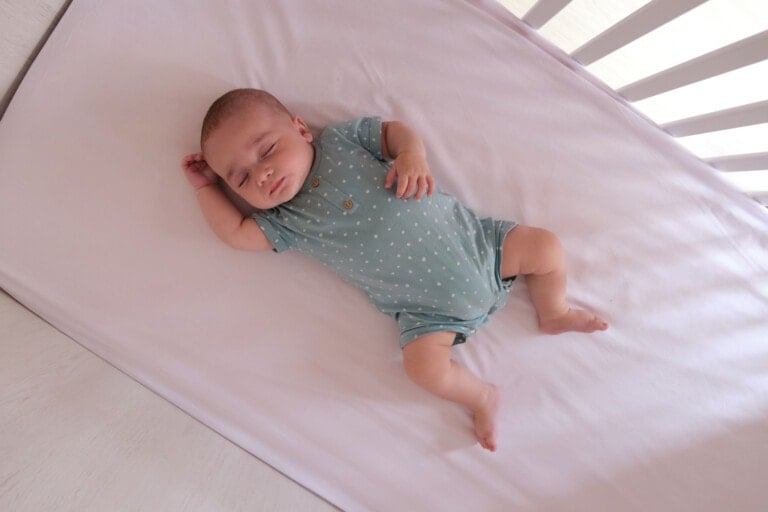 A baby is lying asleep in a crib, following safe sleep guidelines, and wearing a teal polka dot onesie. The baby has one arm raised above their head and is resting on a white mattress with a white crib railing visible on the side.