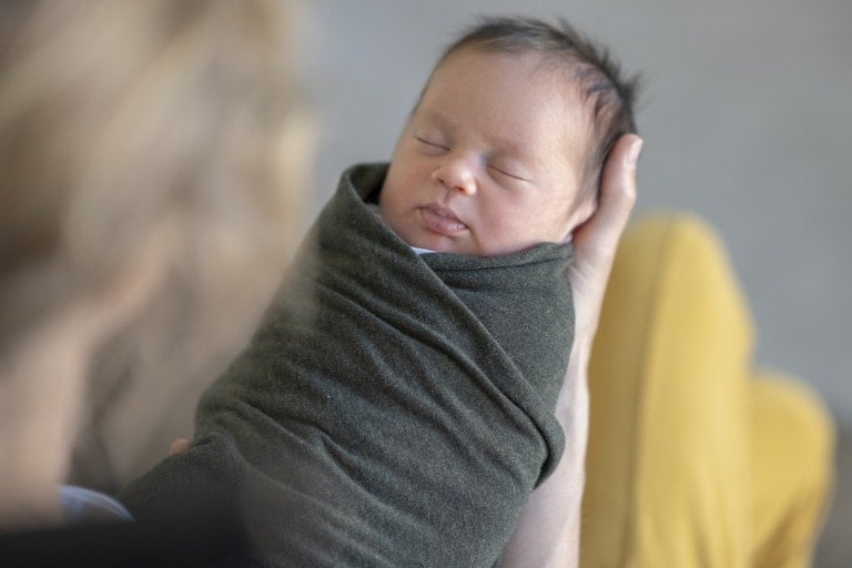 A newborn baby, wrapped in a dark green blanket, is cradled in someone's arms. The baby is sleeping peacefully. The background is blurred, making the baby the focal point of the image—a serene moment before parents transition babies out of swaddles.