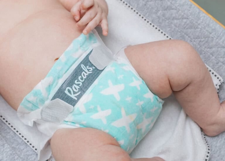 A close-up image of a baby lying on a changing table, wearing a green and white Rascals diaper with a pattern of airplanes. The Rascals Premium Diapers brand is visible on the waistband. The babys arms and legs are partially shown.