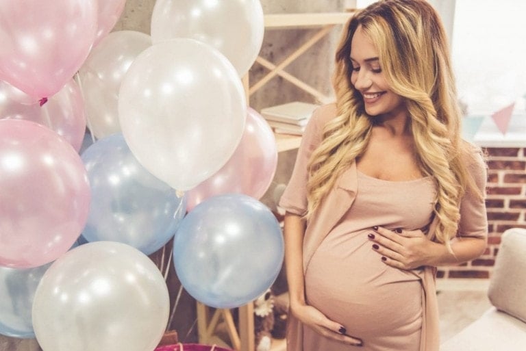 A pregnant woman with long blonde hair, wearing a light pink dress, is smiling and holding her belly. She is standing next to a cluster of pink, blue, and white balloons in a decorated room, where a beautiful baby shower gift basket sits nearby.