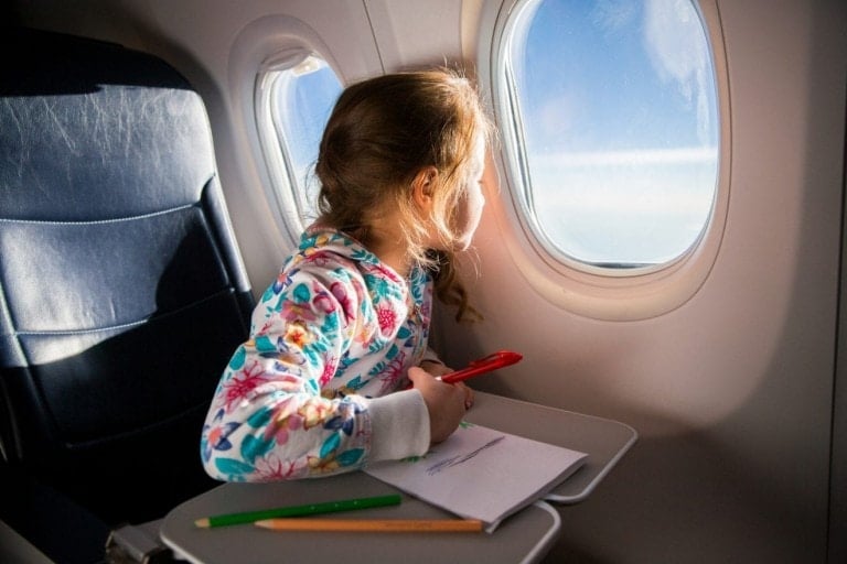 A young girl with long hair wearing a floral hoodie is sitting in an airplane seat, looking out the window during a plane ride with kids. She has a red marker in her hand and colored pencils and paper on the tray table in front of her. Sunlight is streaming in through the window.
