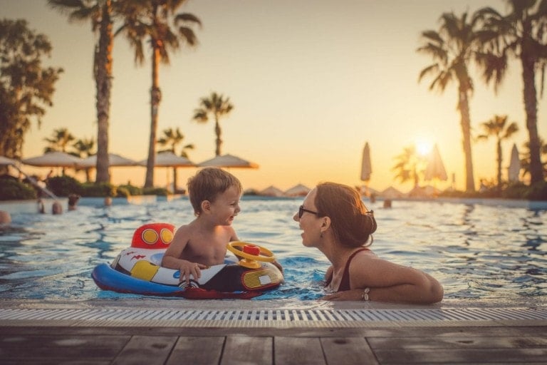 A young child sits in an inflatable boat in a swimming pool, smiling at an adult woman wearing glasses who is partially submerged in the water. Palm trees, lounge chairs, and umbrellas surround the pool area at sunset. Other people can be seen in the background enjoying their Spring Break products for kids.