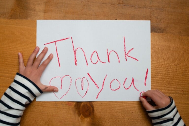 A piece of white paper on a wooden table showcases the words Thank you! written in red crayon. Two hearts are drawn next to the text. A child with black and white striped sleeves is holding the paper and a crayon, creating a delightful example of gratitude art projects.