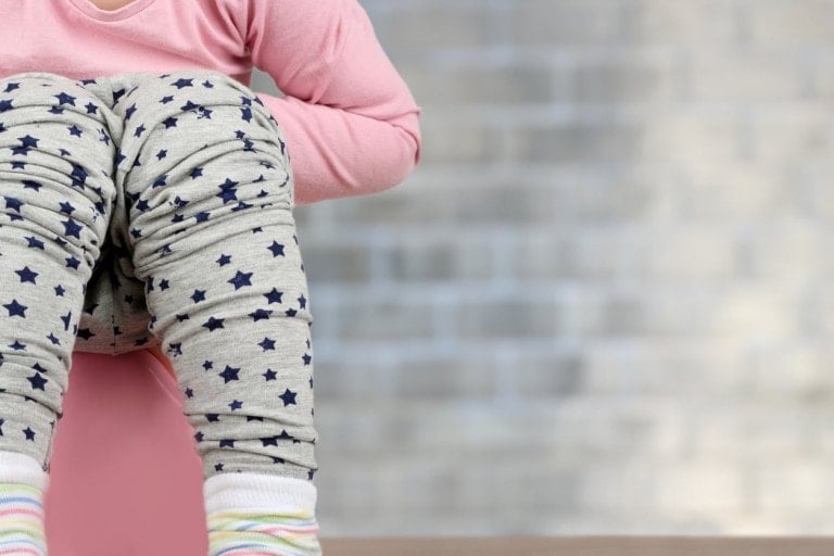 A child wearing a pink shirt, gray pants with navy blue stars, and striped socks is sitting on a pink object. The background consists of a gray brick wall. This could easily be a scene from an article about how to potty train a girl, given the child's concentration and surroundings.