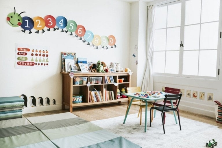 A bright and tidy playroom, showcasing excellent playroom organization, features a wall with colorful numeric caterpillar wall decals (1-10), a bookshelf stocked with books and toys, a small table with two chairs, and various educational posters. Light pours in through a large window, illuminating the space.