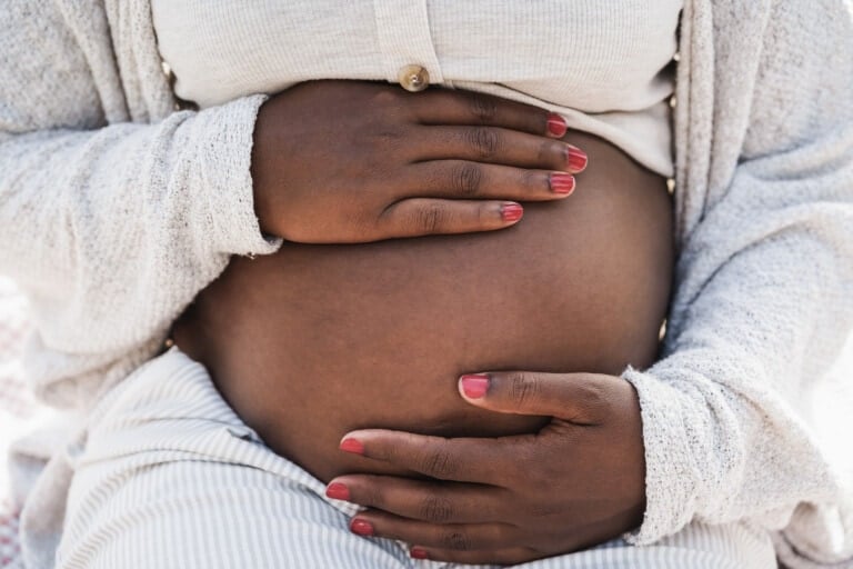 A close-up of a pregnant person's belly with both hands resting on it. The person is wearing a light-colored top and a cardigan, and has their fingernails painted red. This serene moment captures the essence of making pregnancy easier with comfort and style.