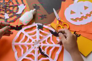 Hands are shown arranging black paper bats on a white paper spiderweb. Scattered around are easy Halloween crafts for kids, including a pumpkin face cutout, paper ghosts, a glue bottle, a pair of scissors, and various colored paper. This scene is full of simple and fun Halloween craft ideas!
