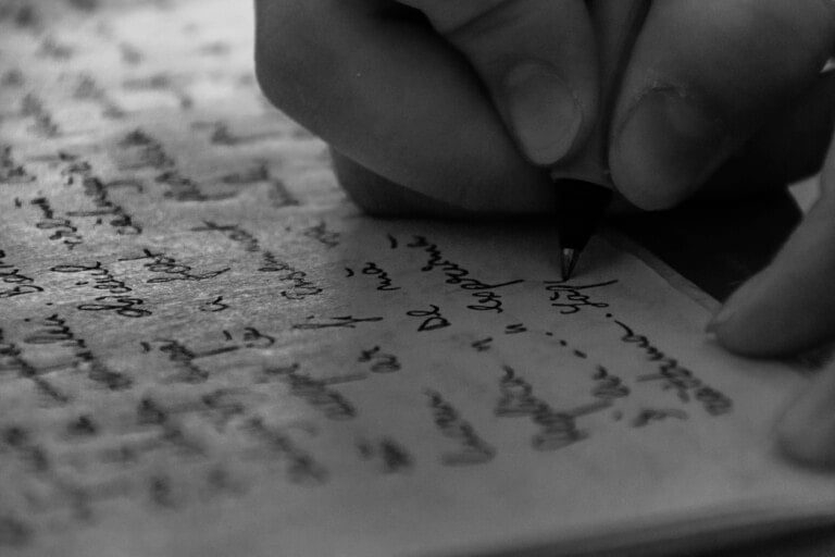 A close-up, black and white image of a hand holding a pen and writing in a notebook. The handwriting is cursive and fills several lines on the page. The image captures the essence of the woman I was before I had kids, focusing on the fingers and the pen's tip touching the paper.