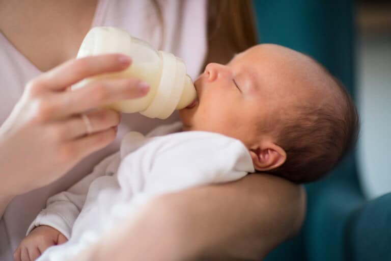 A baby in a white outfit is cradled in someones arms, contentedly feeding from a bottle. The adult, dressed in a light-colored top, fosters a calm and cozy environment, showcasing the serene balance of combo feeding with both breastfeeding and formula.