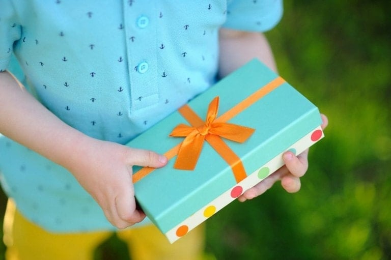 A child wearing a light blue shirt with small anchor patterns holds a rectangular, teal gift box with an orange ribbon and bow. The edges of the box feature a colorful dotted pattern. Opting for gifts that aren't toys, the scene is set against a grassy, green background.