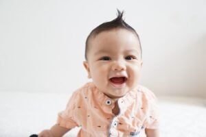 A baby wearing a light beige shirt is smiling widely with an open mouth, showing a small part of their teeth. The baby, whose parents are considering boy names that start with K, has a short tuft of hair standing up on top of their head. The background is a plain white wall.
