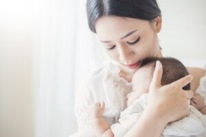A woman with dark hair gently holds and looks down at an infant against her chest. Bathed in soft natural light from the left side of the frame, this peaceful and intimate moment evokes the essence of Korean names, reflecting a timeless bond.