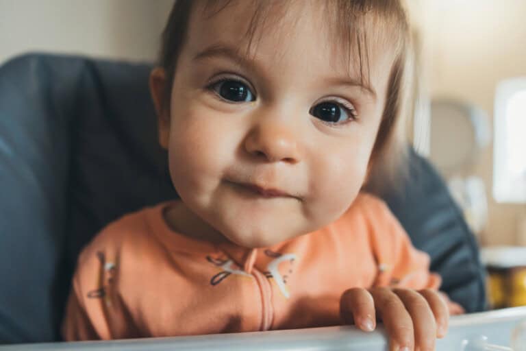 A close-up image of an 18-month-old girl with wide eyes and a slight smile, wearing an orange outfit. The baby is holding onto the edge of a highchair and looking directly at the camera. The background is softly blurred.