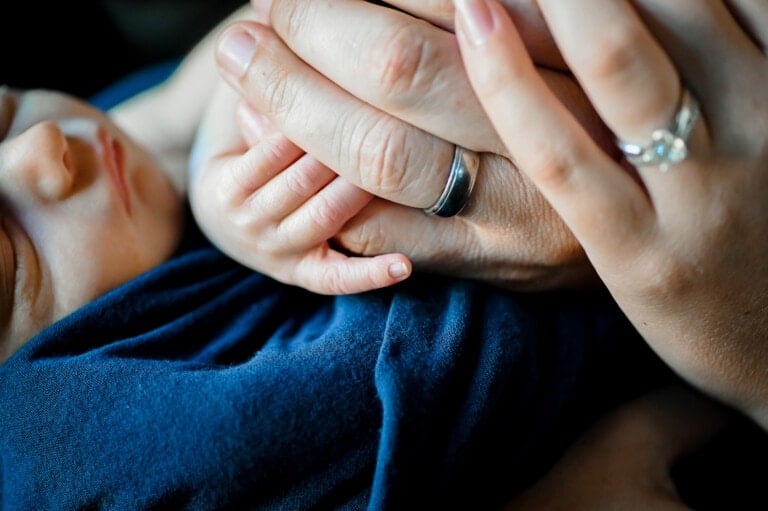 A close-up image of a newborn baby held by an adult. The baby's hand is grasping the adult's finger, capturing the purity of new life and how marriage feels different now. The adult's hand displays a wedding ring and a ring with a stone on another finger. The baby is wrapped in a blue blanket.