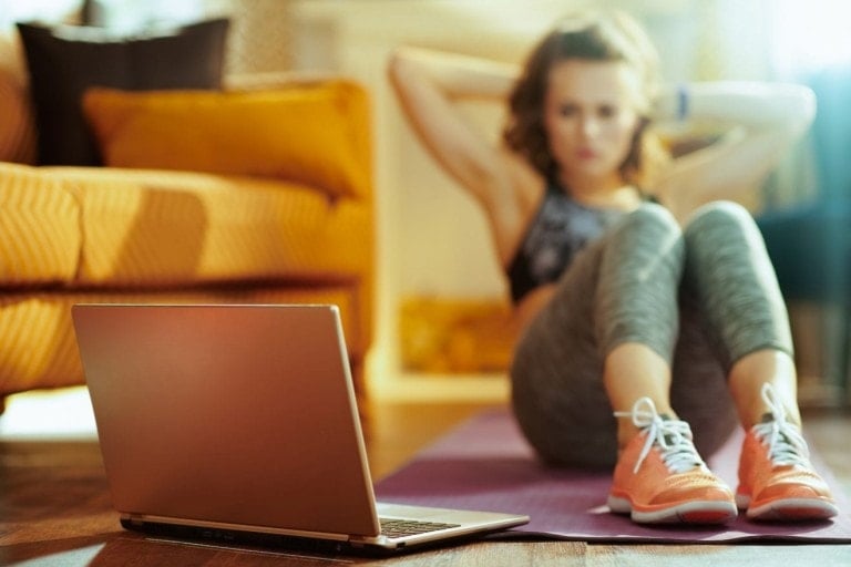 A woman in athletic attire performs abdominal exercises on a purple yoga mat while watching something on her laptop. She is inside a living room, with a sofa and pillows in the background, embodying the spirit of at-home fitness.