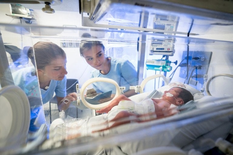 Two medical professionals in blue scrubs are observing a newborn in an incubator. The baby, wearing a diaper and monitored by a tube, is surrounded by medical equipment. The setting clearly appears to be the NICU, emphasizing the high level of care provided for these infants.