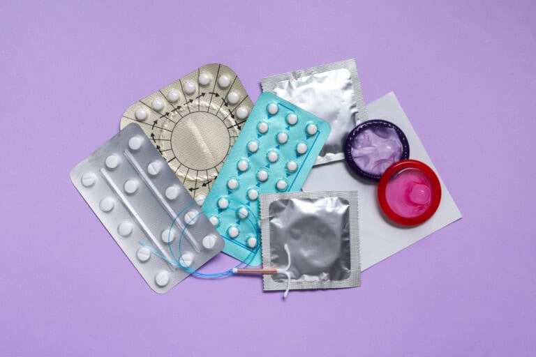 A variety of contraceptives are displayed on a purple background, perfect for those navigating birth control options. Included are blister packs of birth control pills, condoms in foil wrappers, and an intrauterine device (IUD).