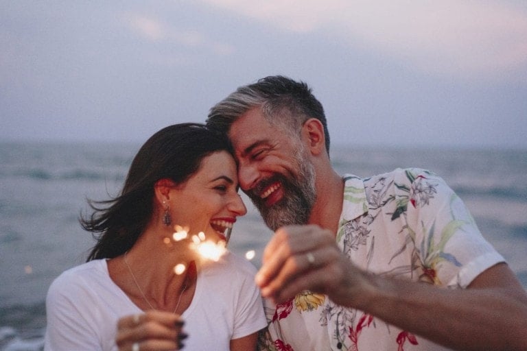 A man and a woman stand on a beach during twilight, smiling and leaning their heads together. They hold sparklers, with the ocean visible in the background. The man has gray hair and a beard, while the woman has long, dark hair—making it a perfect summer date night scene.