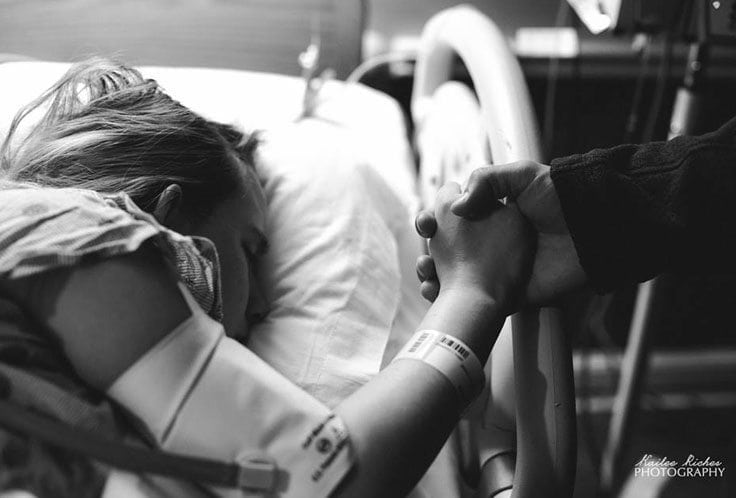A black and white photo shows a person lying in a hospital bed, possibly in active labor, holding hands with another individual standing beside the bed. The person in the bed is wearing a hospital gown and wristbands, seemingly resting or asleep.