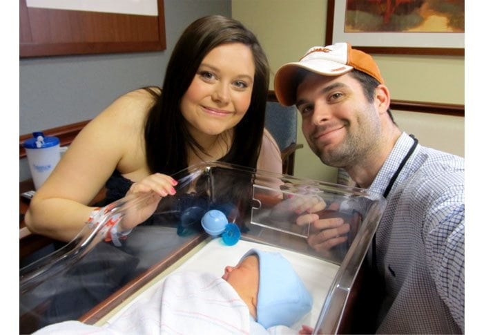 A woman and a man smile at the camera while standing next to a baby in a hospital bassinet, capturing their birth story. The baby is swaddled in a blanket and wearing a blue hat. The man, in his baseball cap and checkered shirt, stands proudly beside them with the hospital room visible in the background.