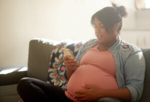 A pregnant woman sits comfortably on a gray couch, holding a smartphone in her right hand and resting her left hand on her belly, perhaps timing labor contractions. She is wearing a pink shirt and a denim jacket with a floral blanket draped over the couch.