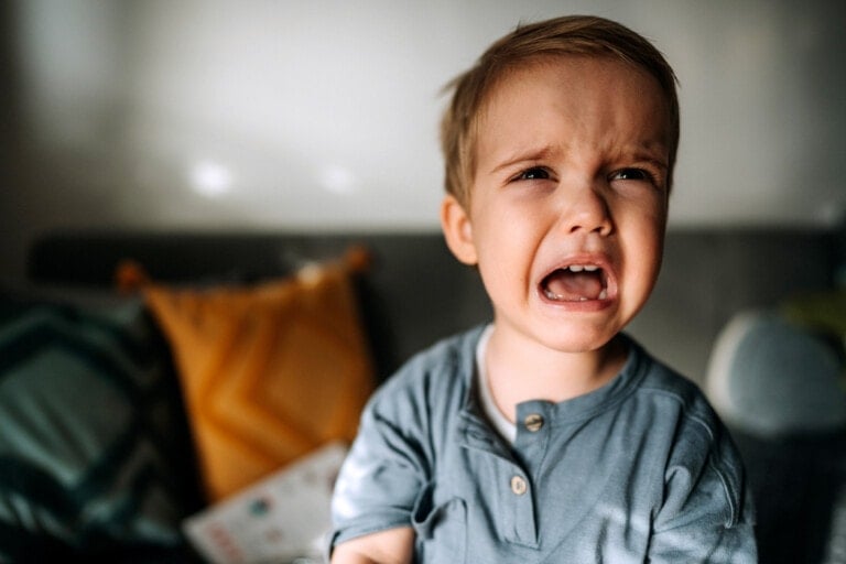 A young child with short brown hair and a blue shirt is crying indoors. The blurred background includes a decorative pillow with a yellow zigzag pattern and a dark-colored couch partially visible. The natural lighting sets the scene often associated with toddler bedtime tantrums, challenging many parents.