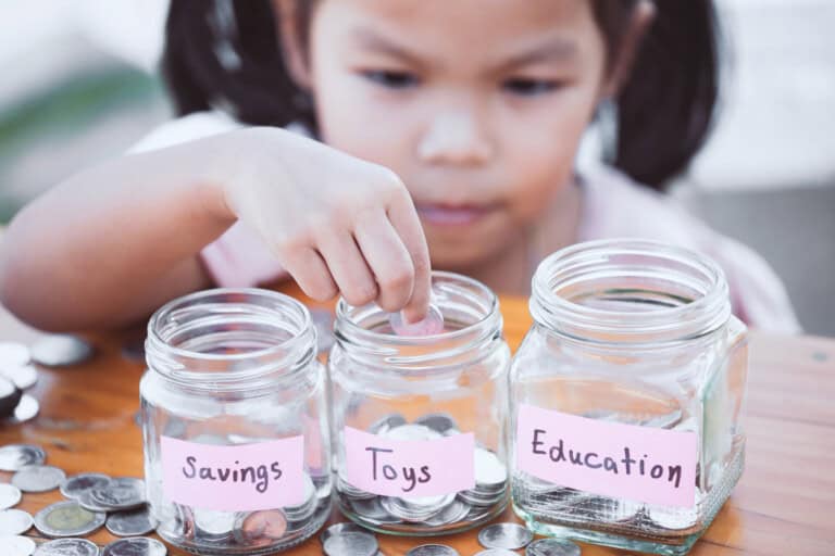 A young child is placing a coin into one of three labeled glass jars. The jars are labeled Savings, Toys, and Education, and each contains coins. Additional coins are scattered on the wooden table around the jars.