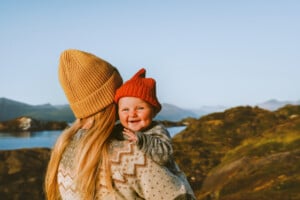 A woman with long blonde hair, wearing a mustard-colored beanie, holds a smiling baby wearing a red beanie. They are outdoors near a body of water and mountains are visible in the background. Both are dressed in warm clothing. The scene evokes the charm of Scandinavian girl names like Astrid or Freja.