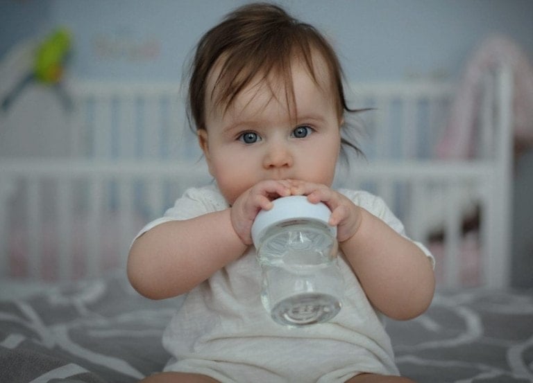 A baby with light brown hair and blue eyes is sitting on a bed, holding and sucking on a clear plastic container with a white lid. In the background, there is a white crib with some toys and a blanket visible; it makes one wonder about the importance of water for baby formula at this stage.