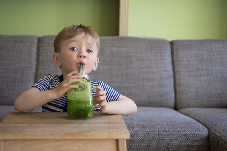 A young child with short brown hair is sitting on a grey couch, sipping a green smoothie through a straw from a mason jar. The child, dressed in a blue and white striped shirt, is seated at a small wooden table in a room with green walls.