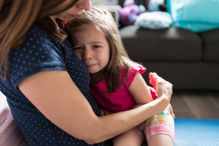 A young girl with long brown hair wearing a pink shirt and multicolored shorts is being comforted by an adult woman with brown hair and a navy blue polka dot shirt. The girl looks upset, possibly due to typical toddler tantrums, as they sit on a blue floor mat in the living room.