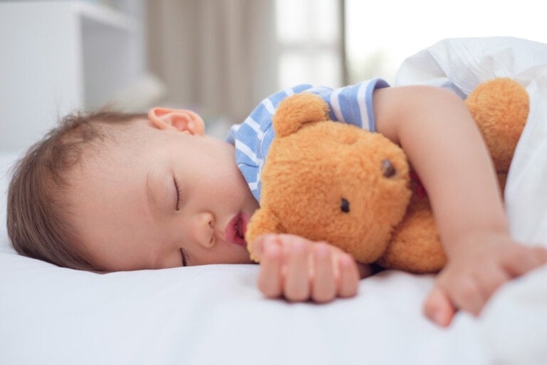 A baby in a blue striped shirt is sleeping on a white bed while cuddling a brown teddy bear, perfectly adhering to their nap schedule. The baby's eyes are closed, and their hand is wrapped around the stuffed toy. The background is softly blurred, indicating an indoor setting.
