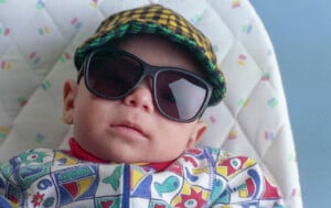 A baby wearing large black sunglasses and a yellow plaid cap is seated in a patterned baby seat. The hipster baby's outfit features colorful abstract shapes on a white background.