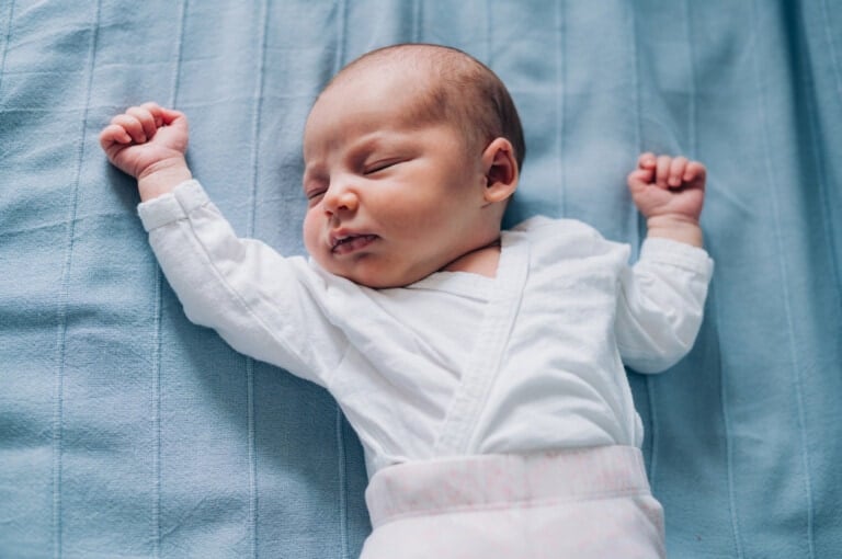 A newborn baby is lying on a blue blanket, wearing a white long-sleeved onesie—perfect to dress baby for safe sleep. The baby is sleeping with both arms raised and hands lightly clenched into fists, with a peaceful expression on their face.