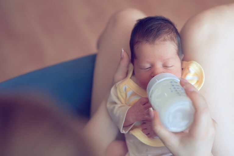 A person is feeding an infant from a bottle while cradling the baby in their lap. Despite the ongoing debate of breastfeeding vs. pumping, the baby, dressed in a yellow outfit, appears to be calmly drinking the milk. The scene suggests a nurturing and intimate moment.