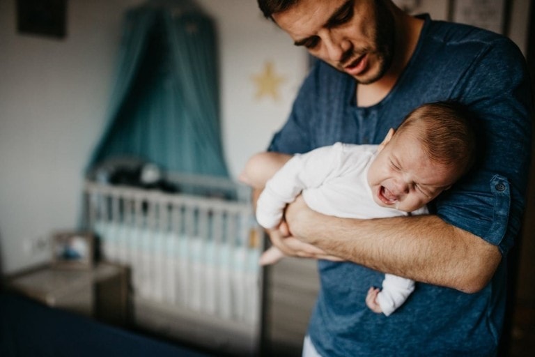 A man in a blue shirt is holding a crying baby dressed in a white outfit. It appears the baby fell off the bed but seems ok. In the background, there is a crib with a canopy and a star decoration on the wall, suggesting the room is a nursery.