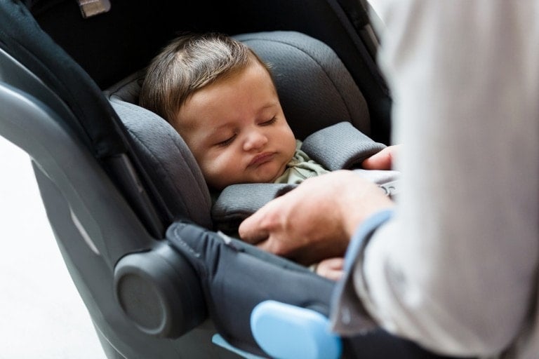 A baby is sleeping in a gray car seat, secured with safety harnesses. An adult's hands are visible, carefully adjusting the harness to avoid common car seat mistakes and ensure the baby is properly strapped in. The scene is indoors and well-lit.