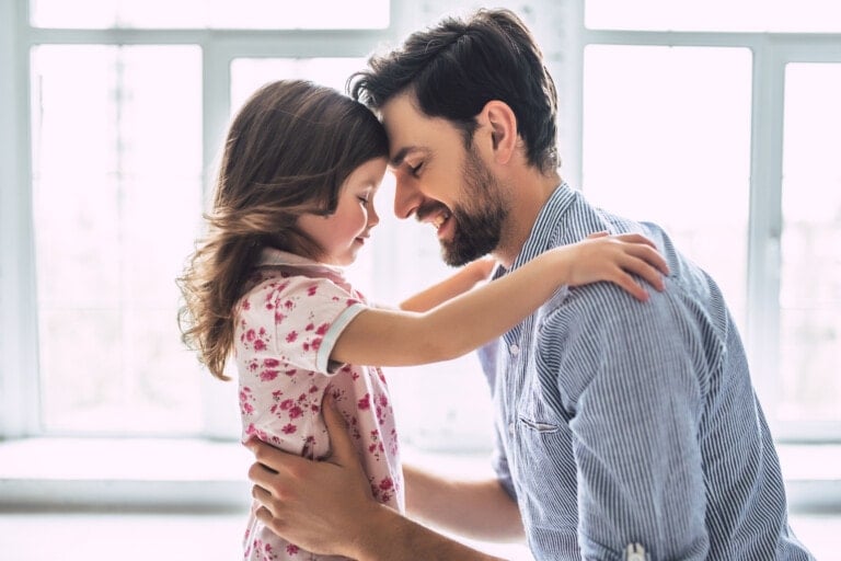 A man and a young girl are facing each other, with their foreheads touching. The man has a beard and is wearing a striped shirt. The girl, in a pink floral dress, has long hair cascading down her back. Inside a bright room with large windows behind them, they share the love that every daughter needs from her father.