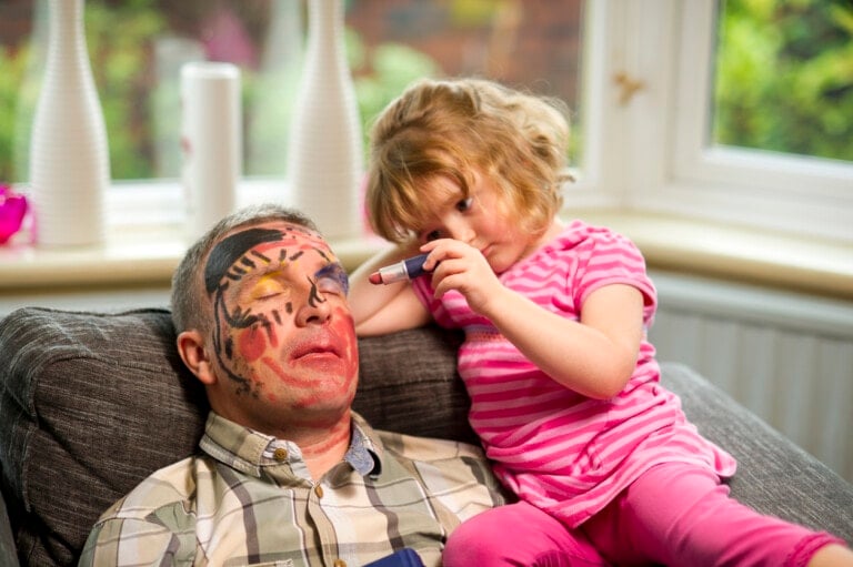 A young girl in a pink striped shirt is drawing on a man's already colorful face with a marker as he sleeps on the couch, enjoying some much-needed self-care. They are in a well-lit room with plants visible outside the windows.