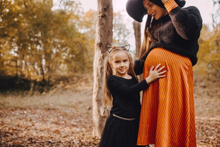 A young girl in a black dress with cat ears stands next to a pregnant woman in an orange striped dress and black hat, touching the woman's belly. They are in a wooded area with fallen leaves on the ground, showcasing creative Halloween costumes for pregnant women amidst nature's autumn splendor.