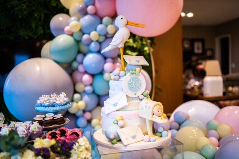 A decorative baby shower cake featuring a stork on top, pastel-colored balloons in the background, and surrounding treats. This exquisite centerpiece is adorned with baby-themed decorations like letters and a small baby figurine, adding to the celebratory atmosphere characteristic of baby shower cakes.