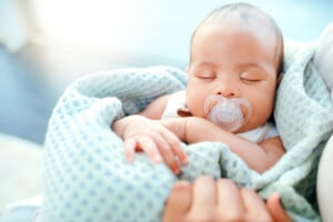 A baby with a pacifier is sleeping, wrapped in a light blue blanket. The baby, perhaps destined for one of those charming boy names that start with "I," is being held, with a hand resting gently next to them. The background is softly lit and slightly blurred.