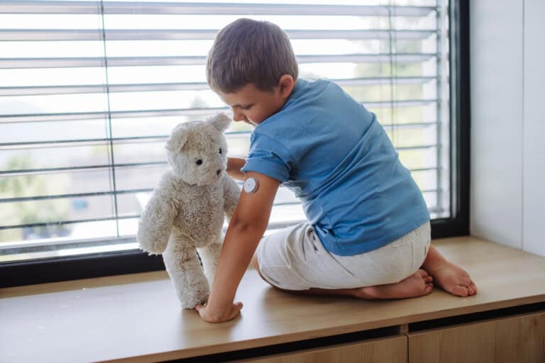 A young boy in a blue shirt and light-colored pants kneels on a wooden bench by a window with blinds. He is placing a stuffed bear in front of him. The boy has a medical sensor on his upper arm due to his type 1 diabetes. The background shows a blurred outdoor view.