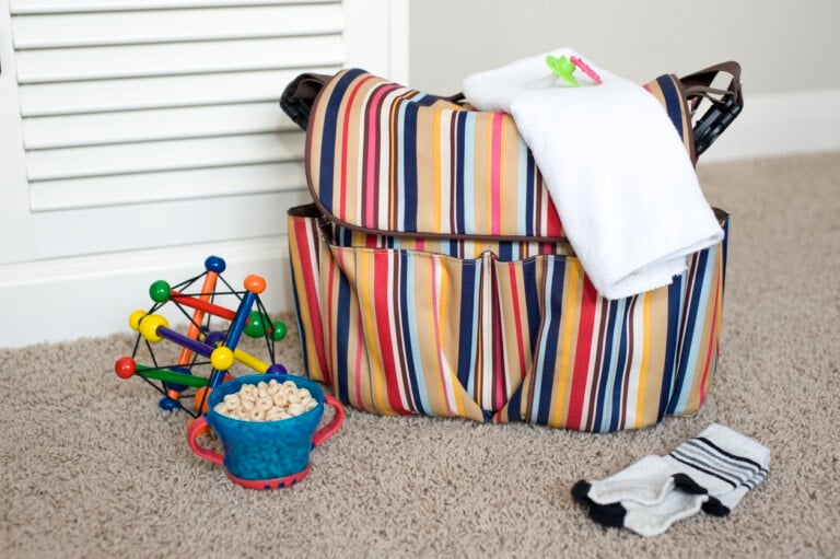 A multicolored striped diaper bag is on a carpeted floor with a white burp cloth draped over it, a multicolored toy, a small blue container of cereal, and a pair of black-and-white socks placed nearby—perfectly packed for any adventure.