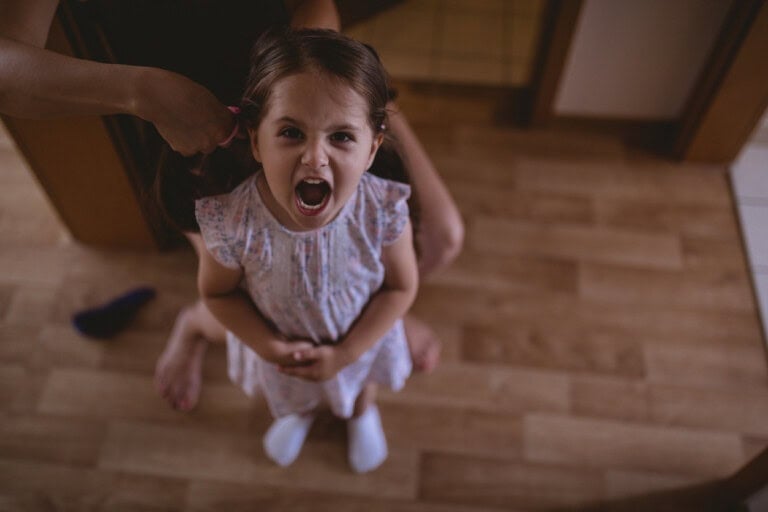 A young girl with a bratty expression stands with her mouth open, seemingly shouting or crying. Her hair is being styled by an adult whose hands are visible, and she is standing on a wooden floor.
