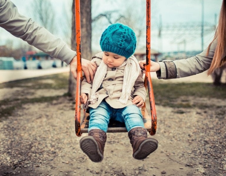 A young child, dressed warmly in a blue knitted hat, beige coat, and jeans, sits on an orange swing. Two adults—embodying a collaborative spirit of parenting with the other woman—stand on either side, steadying the swing with their hands. The background includes trees and a fenced area.