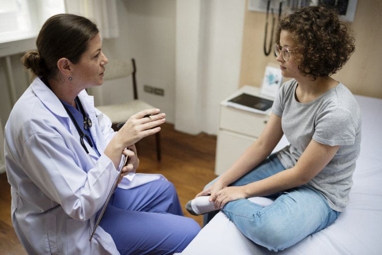 A doctor and a patient are sitting and talking in a medical office. The doctor, wearing a white coat and holding a clipboard, is gesturing with one hand. The patient, dressed in a gray t-shirt and jeans, is sitting on an examination table, listening attentively during her first prenatal visit.