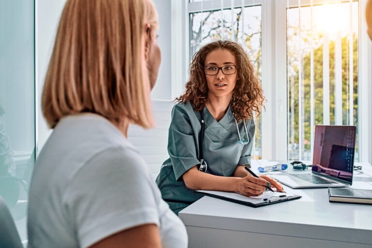 A medical professional wearing scrubs and a stethoscope is seated at a desk, speaking to a woman with blonde hair. The pelvic floor therapist is writing on a clipboard while facing the woman. A laptop is open on the desk, and a window with vertical blinds is in the background.